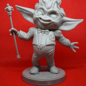 May include: A gray figurine of a whimsical creature with large ears, a mischievous grin, and a flower on its chest. The creature is wearing a tuxedo and holding a jester's staff. The figurine is standing on a round base.