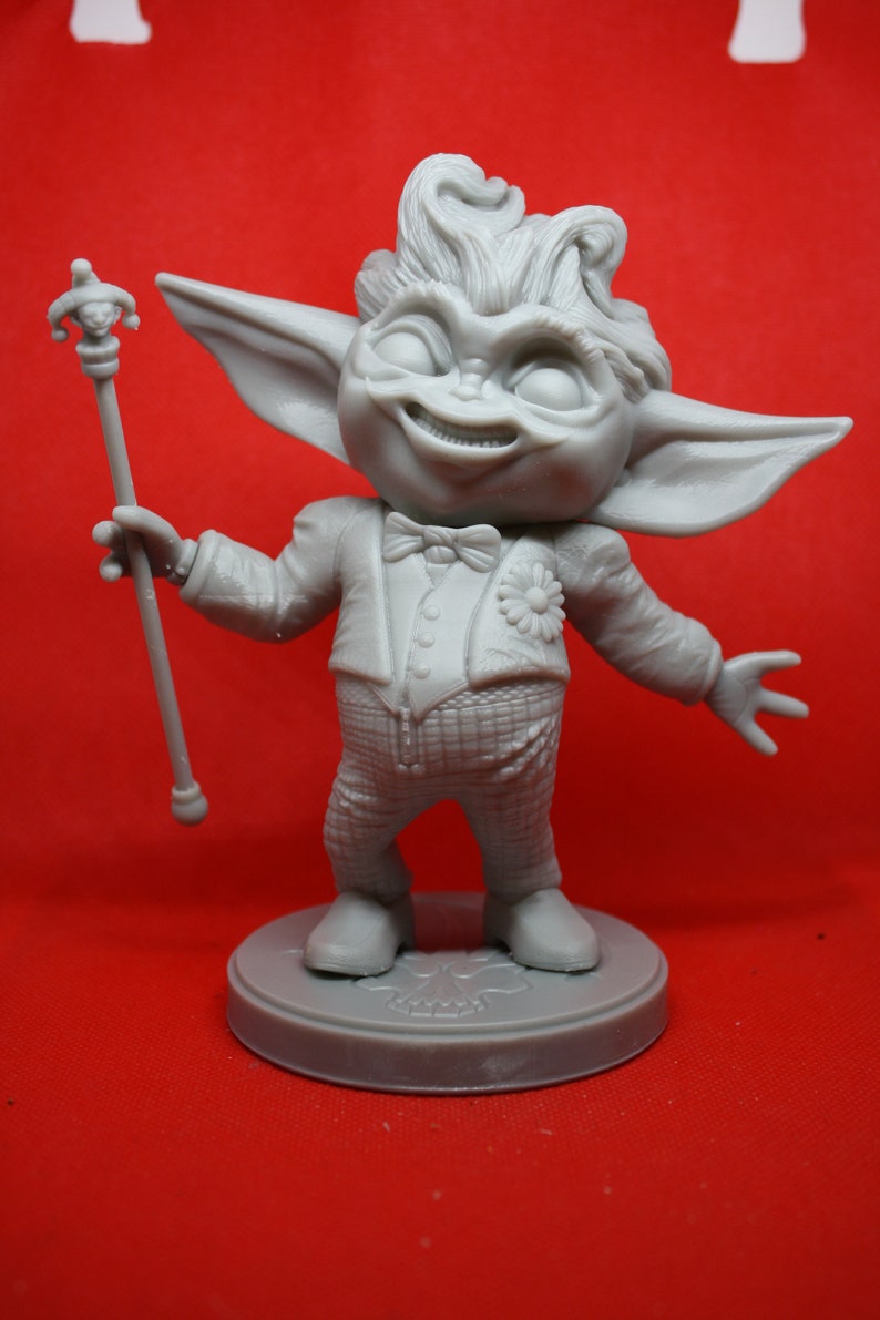May include: A gray figurine of a whimsical creature with large ears, a mischievous grin, and a flower on its chest. The creature is wearing a tuxedo and holding a jester's staff. The figurine is standing on a round base.