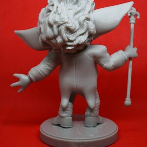 May include: A gray, detailed figurine of a character with a large head of hair, wearing a long-sleeved jacket and pants, holding a staff with a decorative top. The figurine is standing on a round base.
