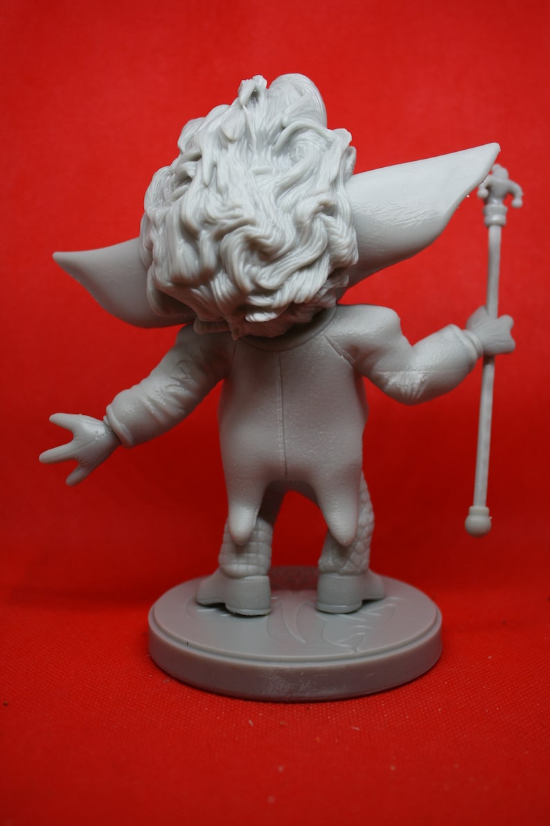 May include: A gray, detailed figurine of a character with a large head of hair, wearing a long-sleeved jacket and pants, holding a staff with a decorative top. The figurine is standing on a round base.