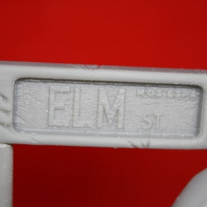 May include: A white sign with the text "ELM ST" in black lettering. The sign is made of a textured material and has a slightly worn appearance.