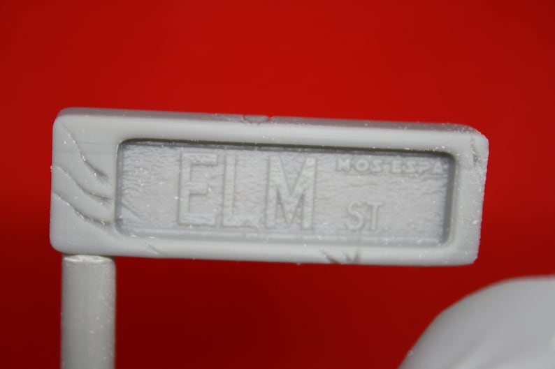 May include: A white sign with the text "ELM ST" in black lettering. The sign is made of a textured material and has a slightly worn appearance.