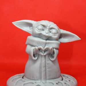 K&ouml;nnte beinhalten: Eine graue 3D-gedruckte Figur eines Charakters aus dem Star Wars-Franchise, bekannt als "The Child" oder "Baby Yoda", die ein Herz mit ihren H&auml;nden formt. Die Figur steht auf einem grauen Sockel mit einem Herzmuster.