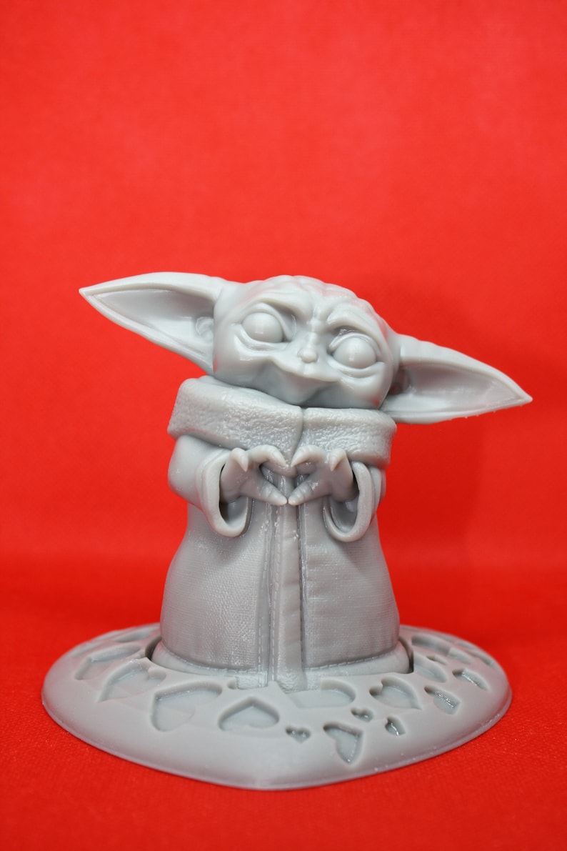 K&ouml;nnte beinhalten: Eine graue 3D-gedruckte Figur eines Charakters aus dem Star Wars-Franchise, bekannt als "The Child" oder "Baby Yoda", die ein Herz mit ihren H&auml;nden formt. Die Figur steht auf einem grauen Sockel mit einem Herzmuster.