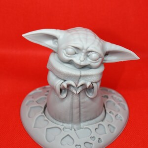 K&ouml;nnte beinhalten: Eine graue 3D-gedruckte Figur eines Charakters aus dem Star Wars-Franchise, bekannt als "The Child" oder "Baby Yoda", die ein Herz mit ihren H&auml;nden formt. Die Figur steht auf einem herzf&ouml;rmigen Sockel mit einem Muster aus kleineren Herzen.