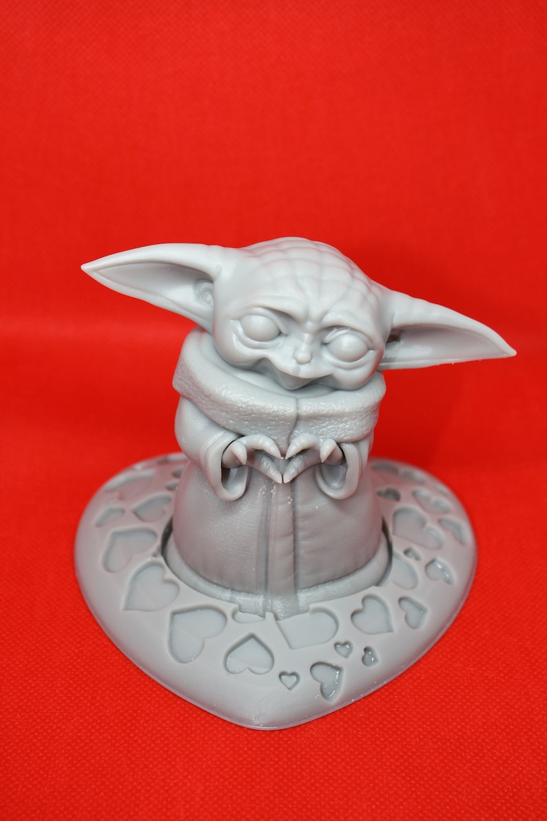 K&ouml;nnte beinhalten: Eine graue 3D-gedruckte Figur eines Charakters aus dem Star Wars-Franchise, bekannt als "The Child" oder "Baby Yoda", die ein Herz mit ihren H&auml;nden formt. Die Figur steht auf einem herzf&ouml;rmigen Sockel mit einem Muster aus kleineren Herzen.