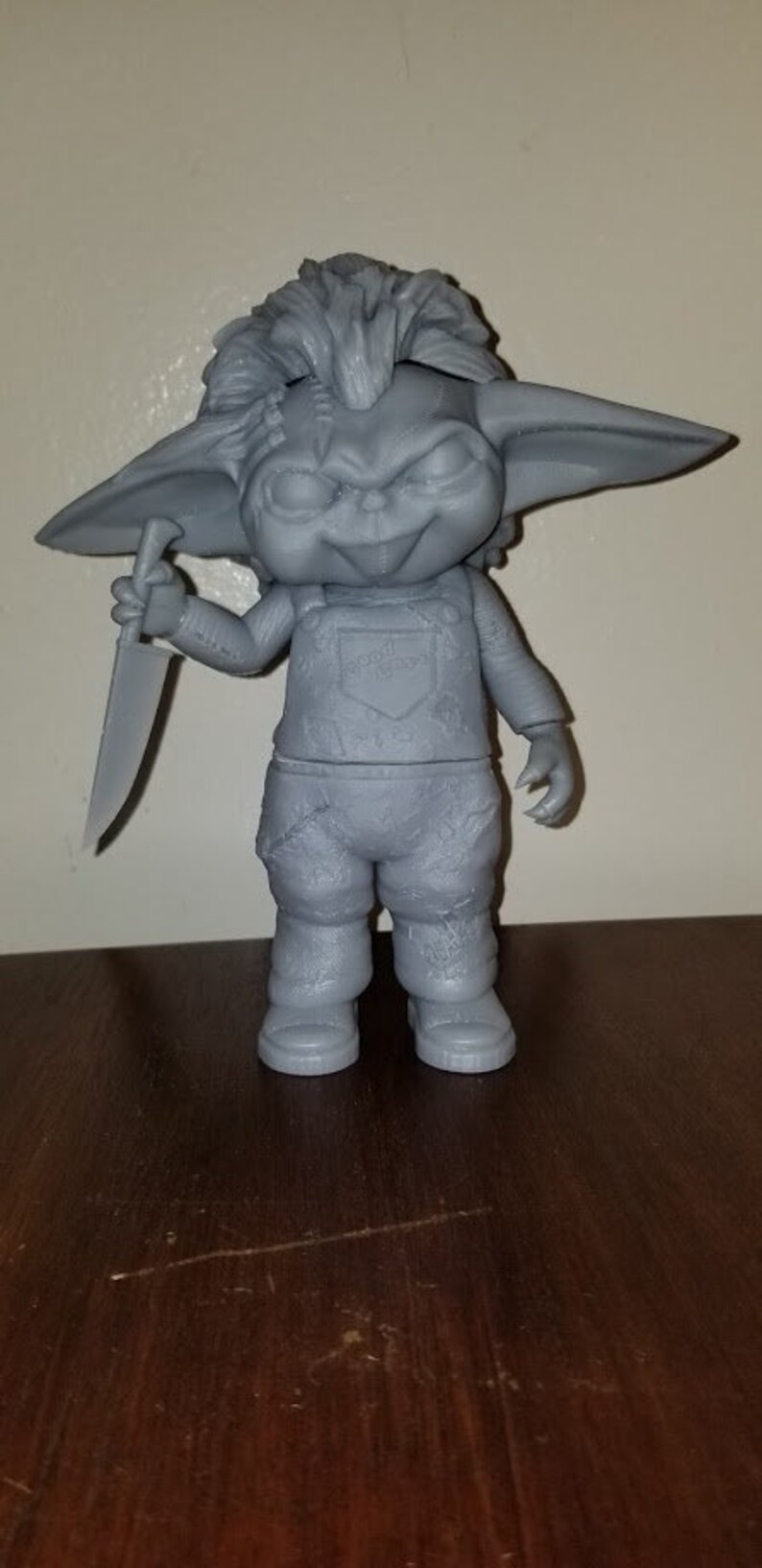 K&ouml;nnte beinhalten: Eine graue 3D-gedruckte Figur eines kleinen Wesens mit gro&szlig;en Ohren, einem verschmitzten Grinsen und einem Messer in der Hand. Das Wesen tr&auml;gt eine Latzhose und hat eine unordentliche Frisur.