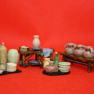 May include: Miniature pottery display featuring various ceramic pieces, including vases, jars, and bowls. The collection includes items in shades of green, beige, and brown, arranged on small wooden benches and trays against a red background.