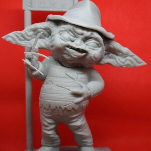 May include: A gray 3D printed figurine of a fictional character wearing a hat and holding a knife. The character is standing on a gray base with the word "Elm St" printed on a sign behind it.