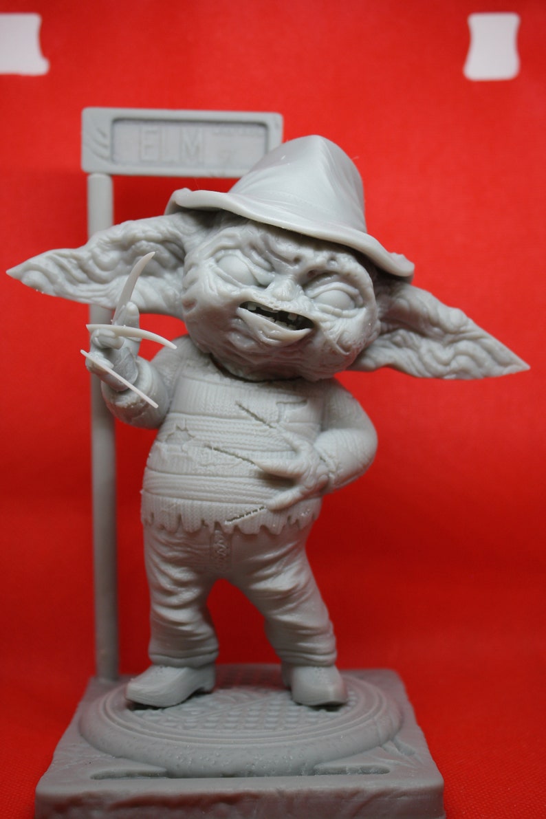 May include: A gray 3D printed figurine of a fictional character wearing a hat and holding a knife. The character is standing on a gray base with the word "Elm St" printed on a sign behind it.