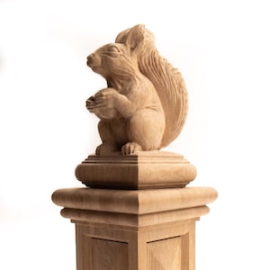 May include: A detailed wooden sculpture of a squirrel holding a nut, mounted on a square pedestal. The squirrel features a bushy tail and textured fur. The pedestal has a layered design, and the sculpture is crafted from light-coloured wood.