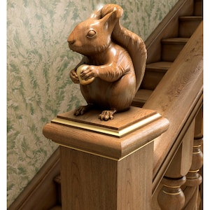 May include: A detailed wooden squirrel sculpture holding a golden nut, perched atop a square wooden post with gold trim. The squirrel is carved with intricate details. The post is part of a wooden staircase.