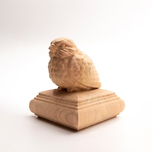 May include: A wooden owl sculpture with detailed feather carvings, perched atop a square, tiered base. The sculpture is made of light-colored wood and has a smooth finish. The owl is depicted in a seated position, with its head turned slightly to the side.