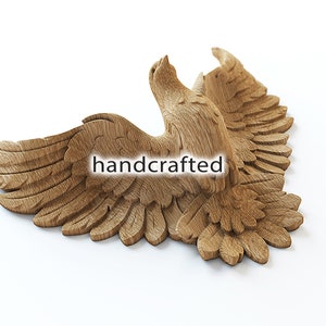 May include: A handcrafted, wooden carving of an eagle in flight. The eagle is depicted with outstretched wings and detailed feathering. The text "handcrafted" is visible on the carving. The carving is a light brown color and is set against a white background.