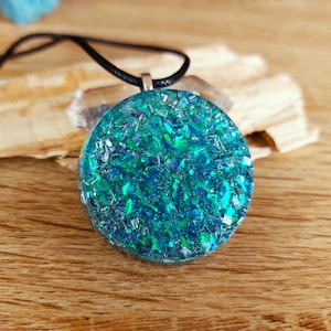 Pode incluir: Colar com pingente redondo com um design azul e turquesa brilhante. O pingente é preenchido com glitter iridescente e suspenso por um cordão preto. O colar é exibido em uma superfície de madeira.