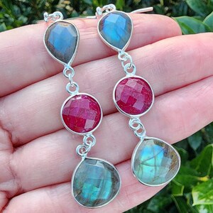 May include: Silver dangle earrings with three gemstone drops. The top drop is a teardrop-shaped blue-green labradorite, the middle drop is a round red ruby, and the bottom drop is a teardrop-shaped blue-green labradorite.