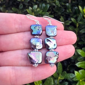 May include: A pair of drop earrings featuring three iridescent, dark purple square-shaped pearls stacked vertically. Each pearl is separated by small silver beads, and the earrings hang from silver-toned metal hooks.