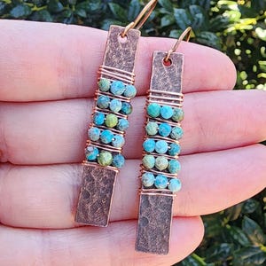 May include: A pair of handcrafted earrings featuring rectangular copper-colored metal pendants. The pendants are adorned with turquoise and green beads, wrapped with copper wire, and have gold-colored ear wires.