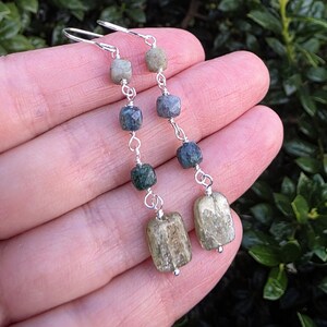 May include: A pair of dangle earrings featuring silver-toned wire links connecting a series of beads. Each earring includes three small, faceted cube-shaped beads in shades of gray and green, ending with a larger, rectangular, textured tan stone.
