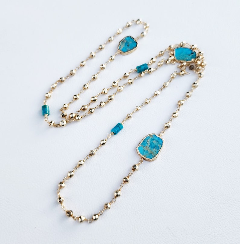 May include: A long gold chain necklace with turquoise stones. The chain is made of small, round beads and features three larger, irregular-shaped turquoise stones.
