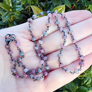 May include: A delicate necklace featuring small, multi-colored gemstone beads in shades of pink, purple, and gray. The beads are strung on a dark, oxidized metal chain with a matching clasp. The necklace is displayed against a natural green background.