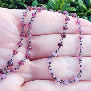 May include: A delicate necklace featuring small, faceted pink gemstones strung on a dark metal chain. The gemstones vary in shade from deep burgundy to pale pink. The necklace is displayed against a blurred green background.