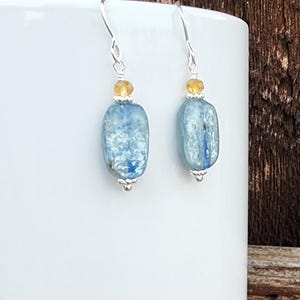 May include: A pair of silver earrings with blue and yellow glass beads. The earrings are hanging from a white surface.
