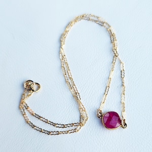 May include: A delicate gold chain necklace with a single, faceted ruby gemstone pendant.