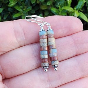 Puede incluir: Un par de pendientes colgantes de plata con un diseño en capas de cuentas de piedra marrón, beige y azul.