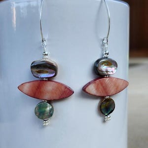May include: A pair of dangle earrings with a silver hook. Each earring features a dark iridescent top bead, a reddish-pink oval bead, and a green iridescent round bead. The earrings are displayed against a white background.