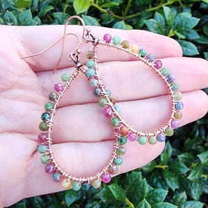 Peut inclure: Boucles d'oreilles en forme de goutte d'eau en fil de cuivre ornées de perles de pierres précieuses multicolores. Les perles sont dans les tons de rose, vert, jaune et bleu. Les boucles d'oreilles sont tenues dans une main.