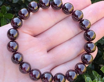 May include: A circular stretch bracelet composed of smooth, polished, deep dark red garnet beads. The beads are uniform in size and shape, featuring a reflective, glossy surface that catches the light, arranged in a continuous loop.