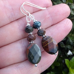Peut inclure: Une paire de boucles d'oreilles pendantes avec des crochets argentés et de petites entretoises argentées. Chaque boucle est composée de trois perles à facettes dans des tons vert foncé, marron et œil-de-tigre, avec une perle ovale en bas.