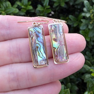 May include: A pair of rectangular drop earrings featuring iridescent abalone shell inlays set within polished gold-tone metal frames. Each earring hangs from a simple gold-tone hook, showcasing natural swirling patterns of green, blue, and purple.