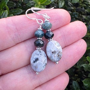 May include: A pair of dangle earrings featuring silver-toned hooks and small silver beads. Each earring includes a round, textured green bead, a faceted black bead, and a larger, smooth, oval-shaped white stone with grey and brown inclusions.