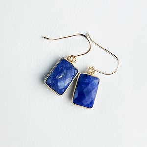 May include: A pair of gold-colored earrings with rectangular, faceted, deep blue gemstones. The gemstones are set in gold-colored bezels and hang from gold-colored ear wires. The earrings are displayed on a white surface.
