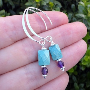 May include: A pair of dangle earrings featuring silver curved ear wires, each holding a faceted rectangular turquoise bead, a small silver spacer, and a single faceted round purple amethyst bead at the bottom, finished with a small silver bead.