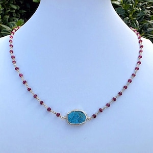 May include: A necklace featuring a central, irregular, bright turquoise stone with a metallic gold-toned edge, suspended on a delicate chain composed of small, faceted, deep red ruby-colored beads linked with thin gold-toned wire segments.