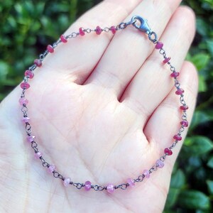 May include: A delicate bracelet featuring small, round, pink and red gemstones strung on a dark metal chain. The bracelet is secured with a silver clasp. The gemstones vary in shade from light pink to deep red.