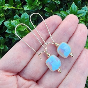 May include: A pair of gold earrings with iridescent white square-shaped pearl-like stones. The earrings have a simple, elegant design.