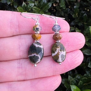 May include: A pair of dangle earrings featuring silver-tone hooks and wire-wrapped construction. Each earring consists of a small faceted bead, a medium brown faceted bead, and a large oval-shaped dark green stone with white inclusions.