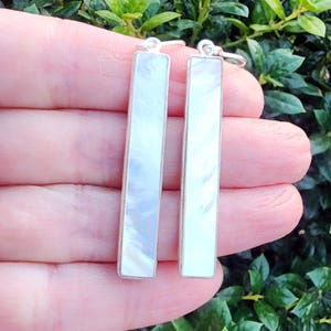 May include: A pair of rectangular earrings with a mother-of-pearl inlay and silver-colored frames. The earrings are shown against a background of green foliage. The earrings are approximately 1 inch in length.