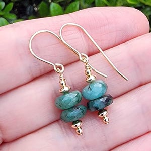 May include: A pair of gold-colored dangle earrings. Each earring features three faceted, teal-colored gemstones and small gold beads. The earrings are suspended from gold-colored ear wires.