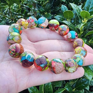 May include: A beaded bracelet featuring colorful, round stones. The stones display a mix of colors including blue, red, yellow, and brown. The bracelet is held in a hand, with a green leafy background.