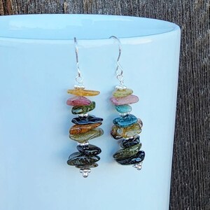 May include: A pair of silver earrings with a stack of colorful, faceted, teardrop-shaped gemstones. The gemstones are layered in a rainbow pattern, with pink, green, blue, and black stones.