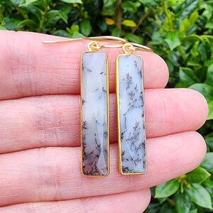 May include: A pair of gold dangle earrings with white and grey dendritic agate stone drops. The stones are rectangular and have a faceted finish.