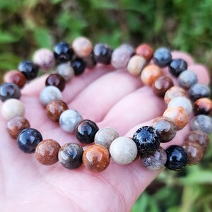 Puede incluir: Un brazalete de cuentas multicolores hecho de coral fosilizado, con tonos de marr&oacute;n, negro, blanco y naranja. El brazalete est&aacute; hecho de cuentas redondas y est&aacute; apilado encima de otro brazalete similar.