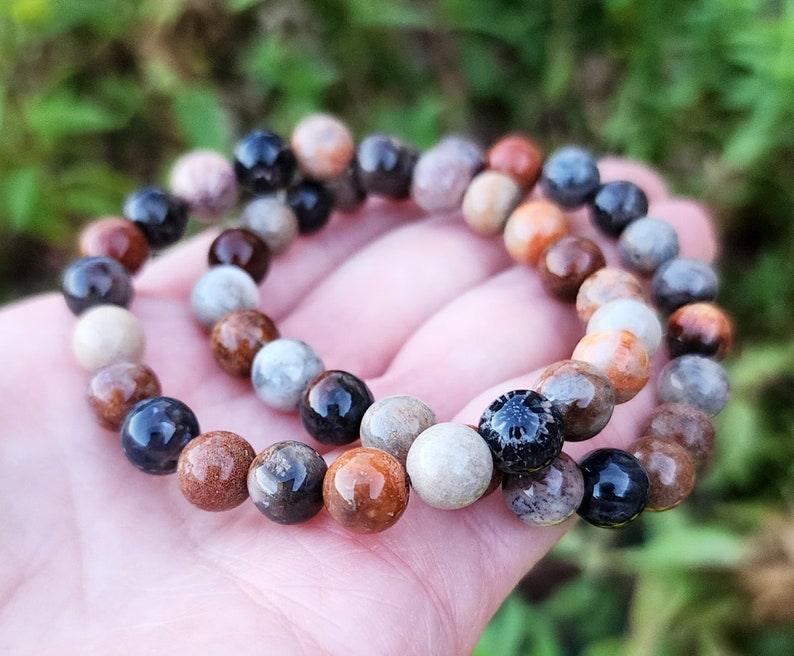 Puede incluir: Un brazalete de cuentas multicolores hecho de coral fosilizado, con tonos de marr&oacute;n, negro, blanco y naranja. El brazalete est&aacute; hecho de cuentas redondas y est&aacute; apilado encima de otro brazalete similar.