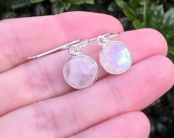 May include: A pair of silver drop earrings featuring round, faceted moonstone gems. Each stone is set in a simple silver bezel frame with a polished finish. The earrings are attached to silver shepherd hook wires, showcasing a subtle iridescent sheen.