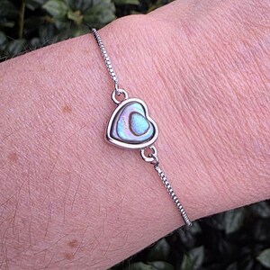 May include: A silver bracelet featuring a heart-shaped pendant with iridescent abalone shell inlay. The bracelet has a delicate chain and is designed to be adjustable. The heart pendant is approximately 0.75 inches wide.